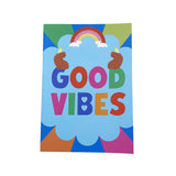 A colourful notebook featuring the words 'GOOD VIBES' in large multicoloured letters inside a light blue cloud shape. Above the text is a rainbow with small clouds, and two brown bear heads sit on either side.