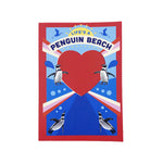 A notebook featuring a red border, a large red heart in the center, penguin illustrations around it, and the text 'LIFE'S A PENGUIN BEACH' on a blue background.