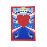 A notebook featuring a red border, a large red heart in the center, penguin illustrations around it, and the text 'LIFE'S A PENGUIN BEACH' on a blue background.