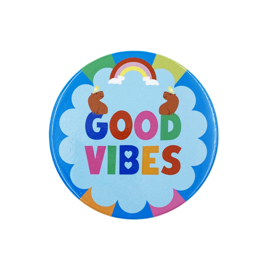 A round coaster featuring the words 'GOOD VIBES' in colourful letters on a light blue cloud, with a rainbow, two brown capybaras