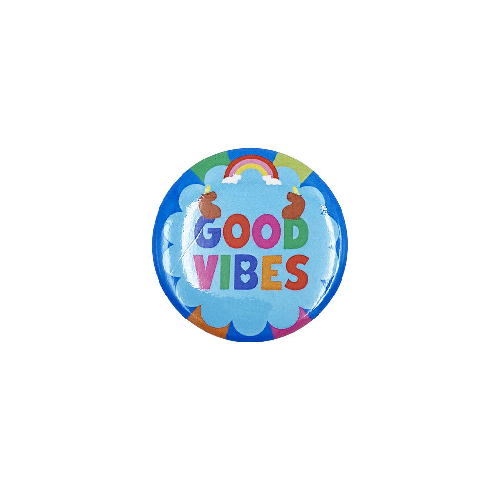 A round pin-back button badge featuring the words 'GOOD VIBES' in colorful block letters on a light blue cloud background, with a small rainbow and sun illustration above, shown on a plain white background.