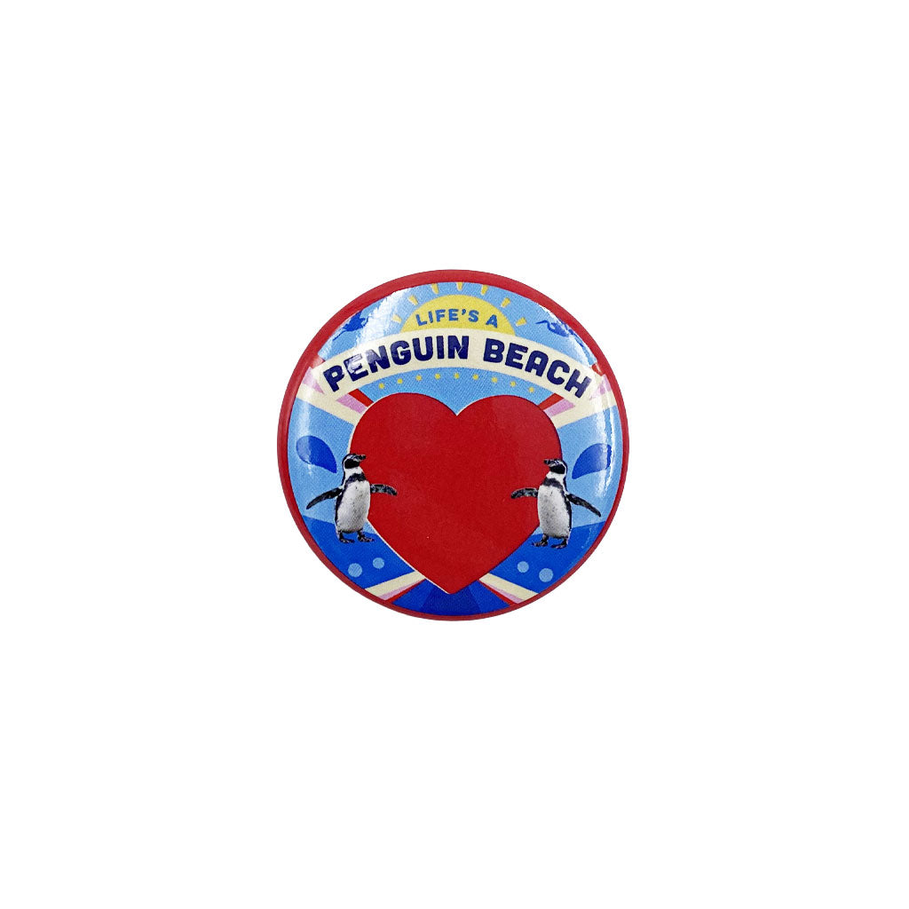 A round button badge featuring a red heart, two penguins, and the text 'LIFE'S A PENGUIN BEACH' on a blue and white background.