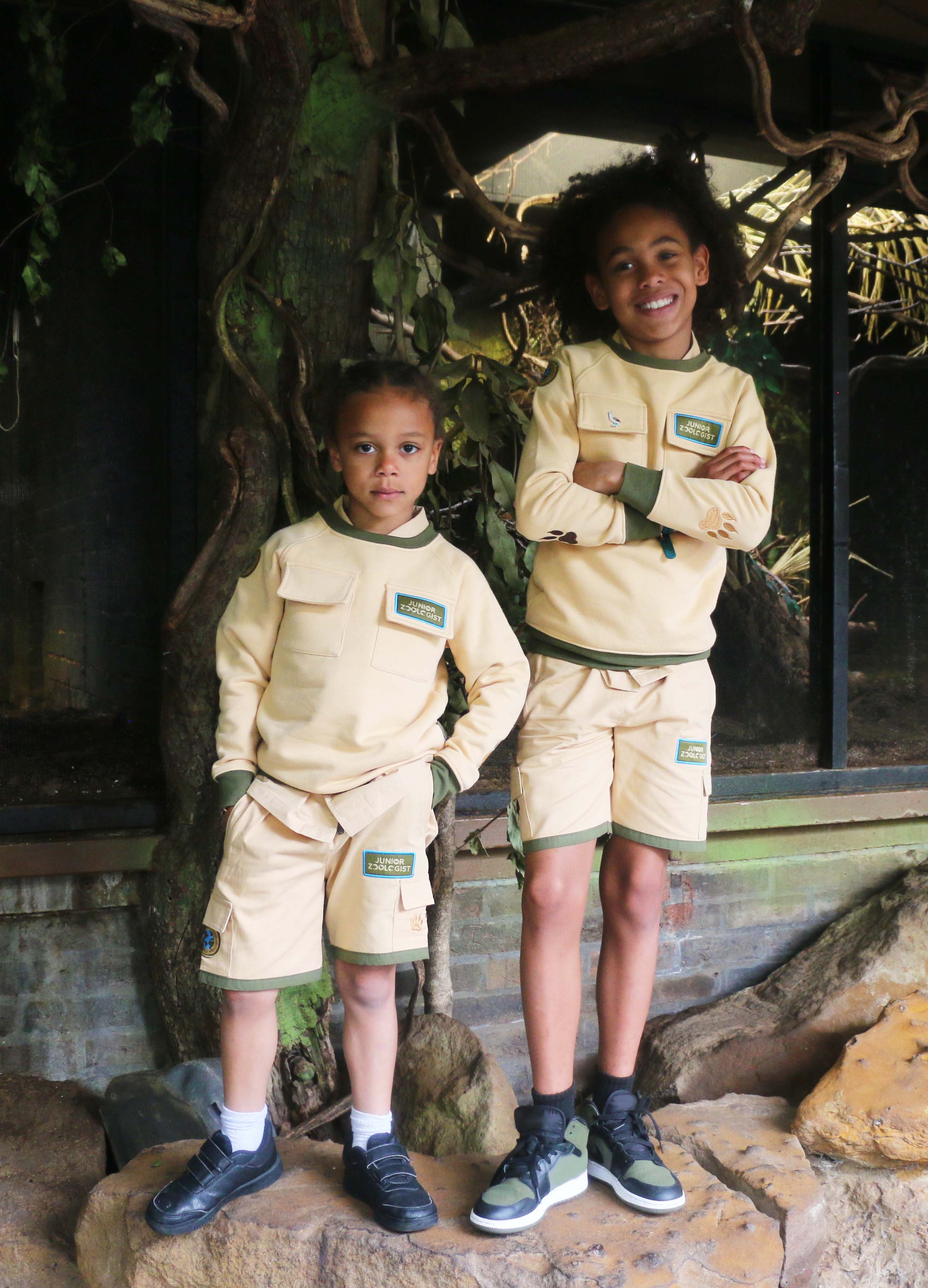 Two children in matching beige uniforms standing in a natural setting with rocks and foliage.